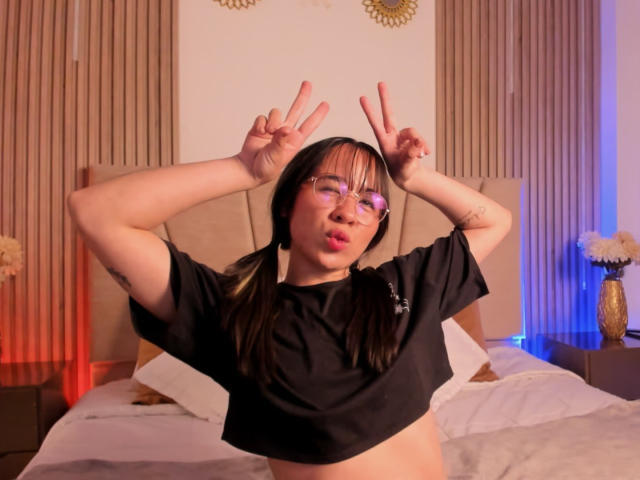 NaomiPearl with pigtails, glasses, and a black crop top, making double peace signs, showing her tits, with a cute, pouty expression in a bedroom.