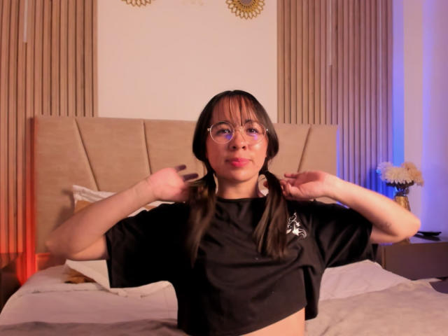 NaomiPearl with pigtails, wearing glasses and a black crop top, pulling at her shirt with her hands, showing her tits, in a softly lit bedroom.