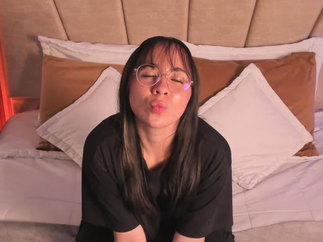 NaomiPearl with black hair, glasses, and puffy lips, pouting, wearing a black shirt, sitting on a bed with white pillows and brown pillows.