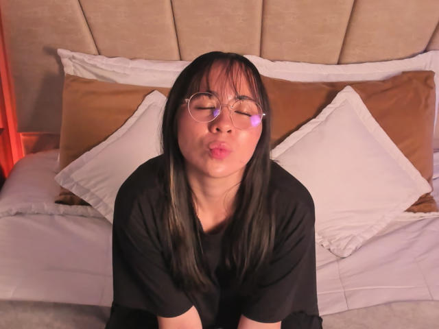NaomiPearl with black hair and glasses, pouting her lips, wearing a black shirt, sitting on a bed with white and brown pillows, in soft red lighting.