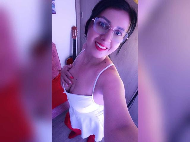 Briananice with glasses, red lipstick, white dress, holding her tits, smiling, black hair, purple-blue light, red floor, guitar in background, selfie, right hand with red nails.