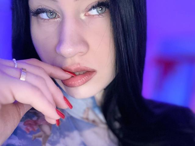 Close-up photo of MaryLori with light skin and blue-green eyes. She has long black hair and is looking slightly to the right. Her red lipstick is slightly parted. She's touching her lips with her right hand, which has red nails and a silver ring. Her eyelashes are long and dark. The blue and purple background creates a contrast with her skin tone. The lighting highlights her facial features and red lipstick. Her expression is subtly seductive.