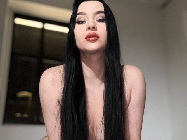 MaryLori has long black hair, pale skin, and red lipstick, looking down with a slight pout. She's topless, showing small tits, and has long, thick eyelashes.