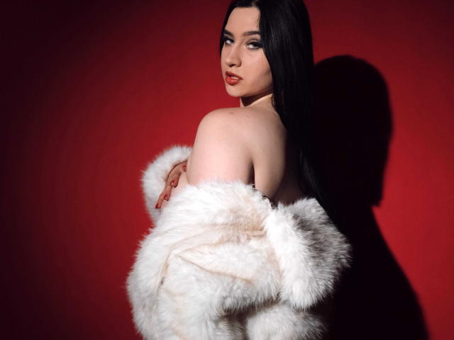 MaryLori with long black hair, red lipstick, and pale skin, wearing a white fur coat, exposing her bare shoulders and back, looking over her shoulder with a sultry expression.