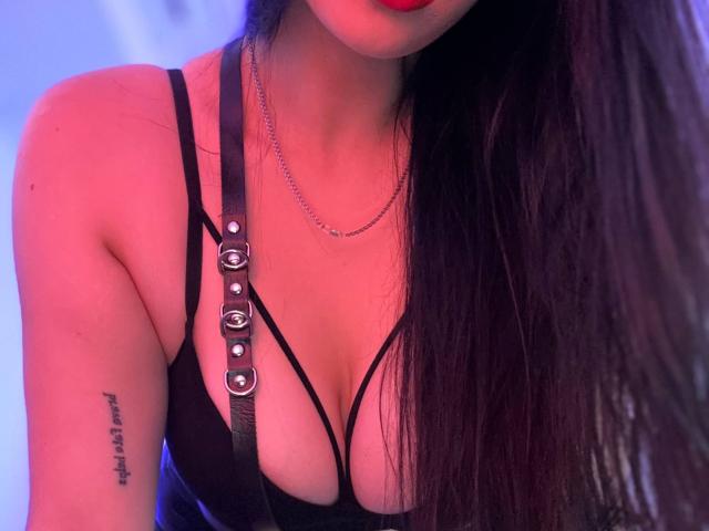 MaryLori, com cabelos longos e pretos, batom vermelho, blusa preta com alças finas que revelam seus seios grandes, colar de corrente prateada e tatuagem no braço direito. Luz roxo-azul..