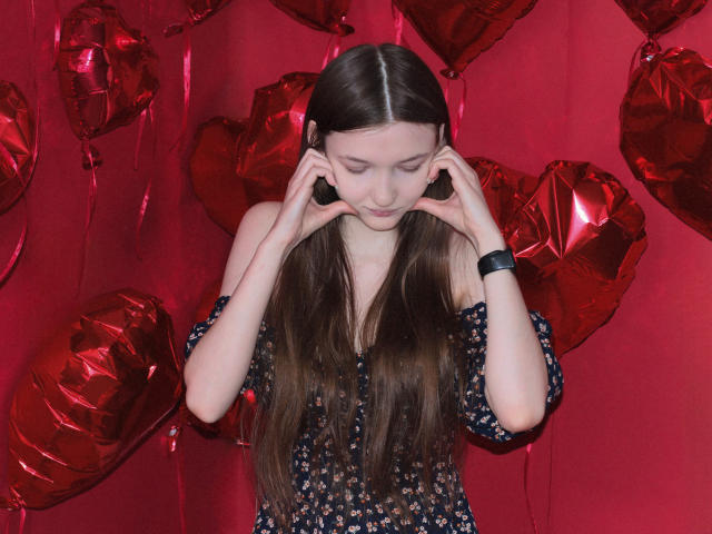 NikaFlorence stands center, with long brown hair, wearing a dark floral off-shoulder top. She holds black heart-shaped headphones around her ears, looking down. Red heart-shaped balloons fill the background, creating a contrast with her dark top. She wears a black wristband on her right wrist. The image has a bright, red backdrop, highlighting the balloons and NikaFlorence's focused expression. The composition is balanced, with the balloons' red color matching NikaFlorence's subtle, dark floral pattern.