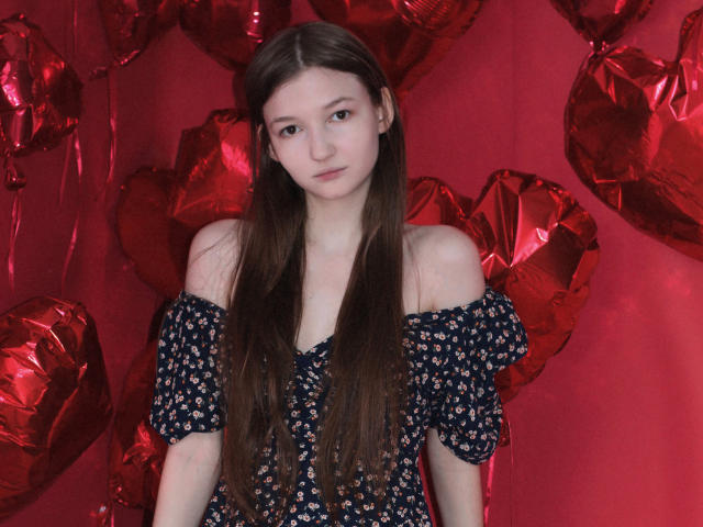 NikaFlorence with long brown hair, off-shoulder blue floral dress, red heart-shaped balloons, and a neutral expression, standing against a red background.