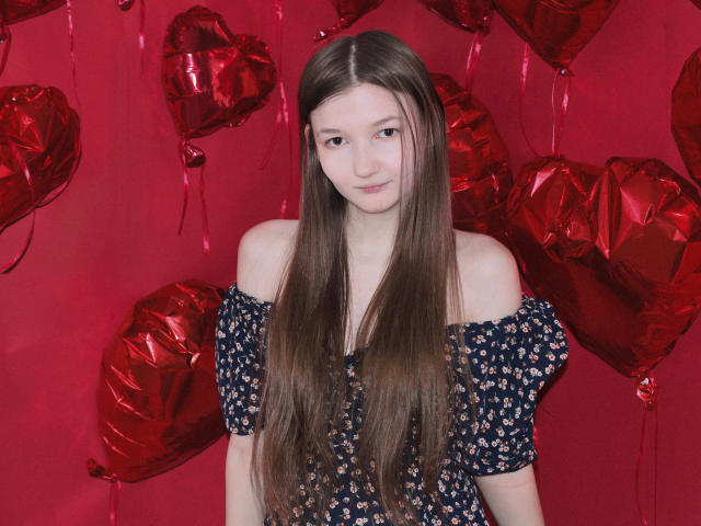 NikaFlorence with long brown hair, wearing an off-shoulder dark blue floral dress, standing in front of red heart-shaped balloons, smiling slightly, looking cute and innocent.