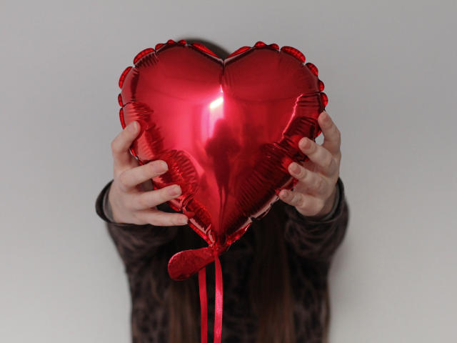 NikaFlorence's hands hold a shiny red heart-shaped balloon, covering her face. Her long brown hair is visible, wearing a dark brown, textured long-sleeve shirt.