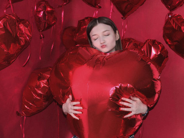 NikaFlorence with red shiny heart balloons, hugging a large red heart, eyes closed, hands gripping the heart, red background, looking relaxed and sweet.