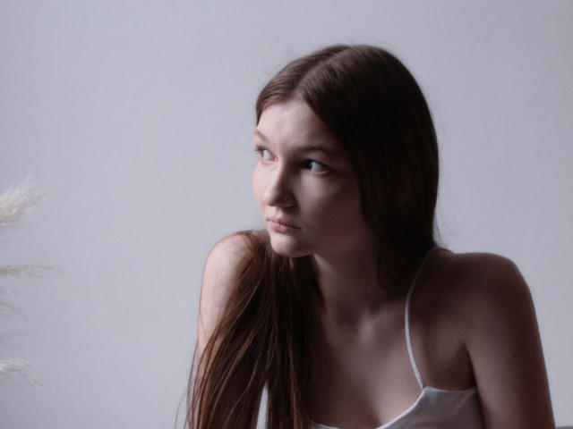 NikaFlorence with long brown hair, wearing a white spaghetti-strap top, looking to the side with a serious expression, her small tits slightly visible, in a soft-lit portrait.