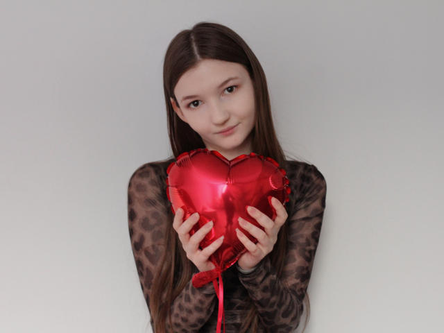 NikaFlorence with long brown hair, holding shiny red heart-shaped balloon, wearing sheer leopard print top, slight smile, holding balloon close to chest with both hands.