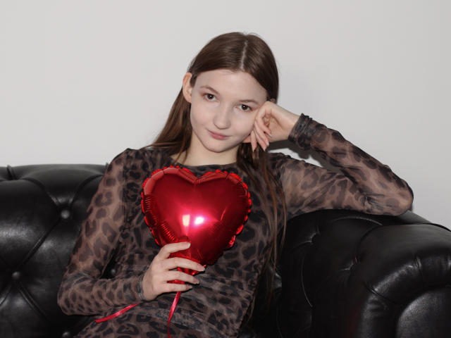 NikaFlorence with brown hair, wearing a sheer leopard print top, holding a shiny red heart-shaped booby, leaning on a black leather couch, looking cute and relaxed.