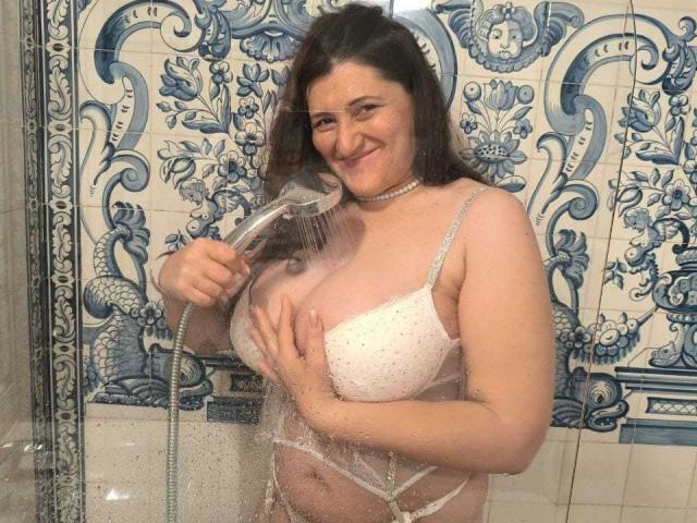 BlissSophia, with wet brown hair, stands in a shower, holding a showerhead to her tits, wearing a white, polka-dot bra and white panties, smiling. Water droplets on glass.