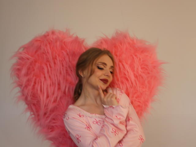 EmilySunshineS with pink fluffy heart, pink long-sleeve pajama top, blonde hair, red lipstick, finger to chin, smiling, soft lighting, fluffy texture, cute, feminine, gentle expression.