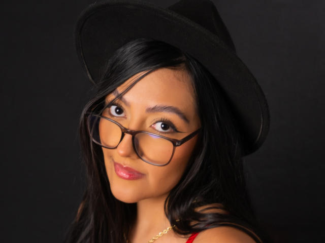 IsabellCollin with black glasses, black hat, and long black hair, showing her cute face and shiny pink lips, looking at the camera with a subtle smile.