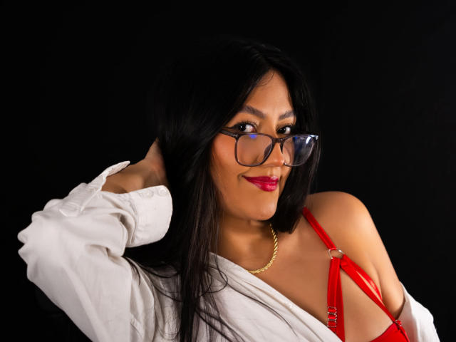 IsabellCollin with long black hair, glasses, red bra, and white shirt, showing her tits, hand in hair, gold necklace, and red lipstick, against a black background.