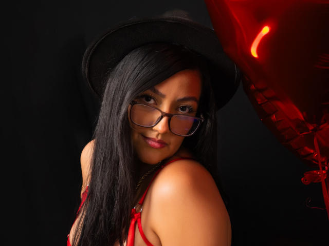 IsabellCollin with long black hair, wearing glasses, a black hat, and red lingerie, looking back with a slight smile, red balloon on the right. Her tits are visible.