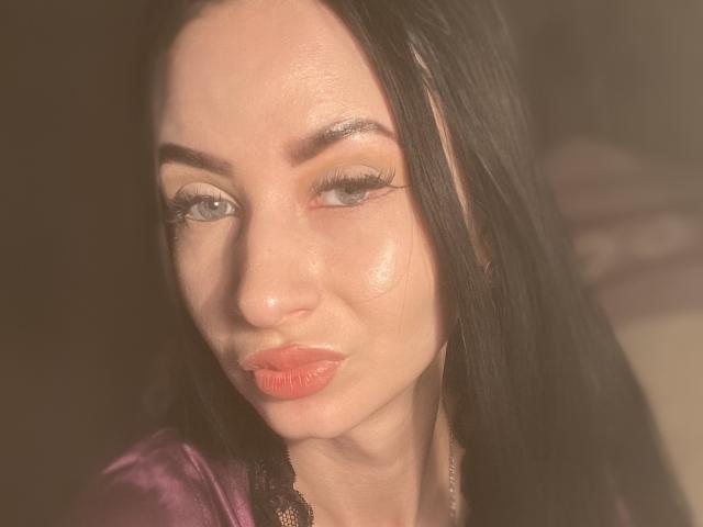 StarrySiren with shiny skin, black hair, and orange lipstick, pouting her glossy red lips, wearing a purple satin top with black lace, has subtle eye makeup and long lashes.