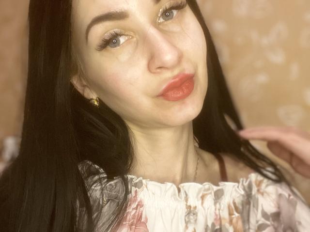 StarrySiren with pale skin, long black hair, blue eyes, red lipstick, floral top, gold earring, pouty lips, slight smile, hand in hair, soft light, cute face.
