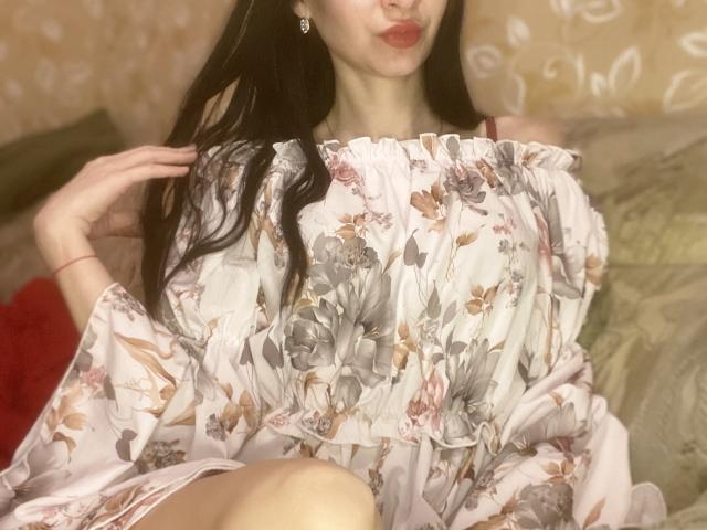 StarrySiren with long black hair, wearing a floral off-shoulder dress, puffy sleeves, and pink lipstick, pouting, hand in hair, sitting with legs crossed, subtle makeup.