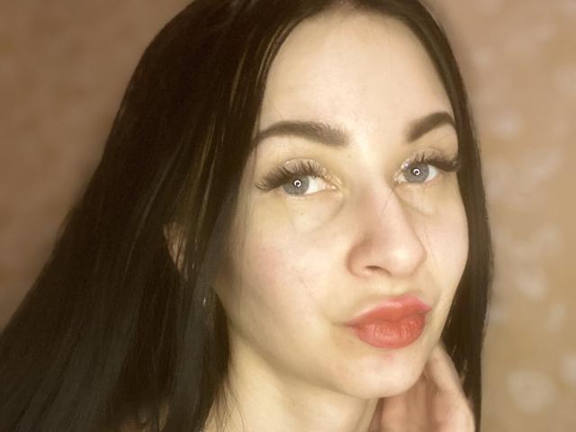 StarrySiren has pale skin, long black hair, blue eyes, and red lips. She's pouting slightly with her right hand touching her neck. Her makeup includes long, thick eyelashes.