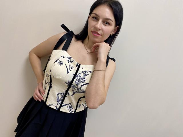 EmmaNoa with black hair, wearing a black and white floral dress, black straps, hand on cheek, smiling, light skin, small earrings, white wall background, standing, looking at camera.