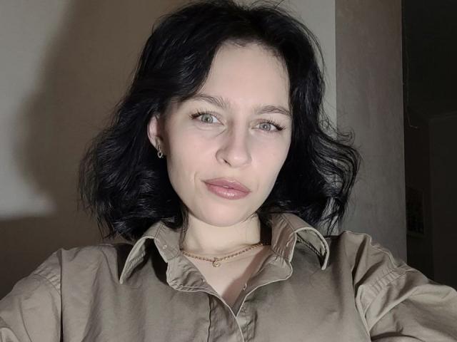 EmmaNoa has pale skin, black wavy hair, green eyes, and a small nose ring. She wears a beige shirt, a gold necklace, and subtle pink lipstick.