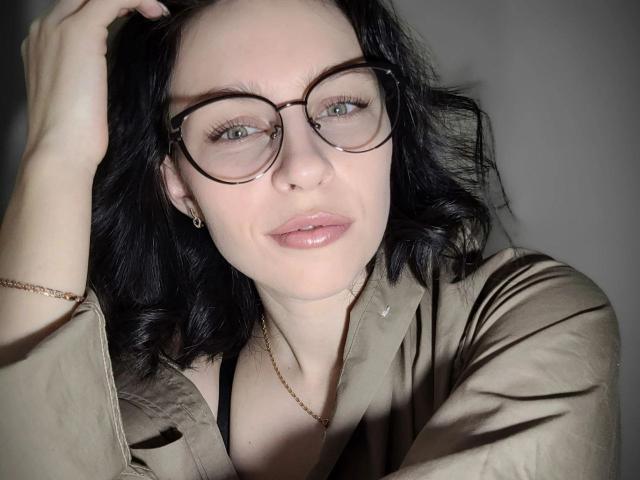 EmmaNoa with black hair, glasses, and glossy pink lips, wearing a beige shirt, black bra visible, hand in hair, subtle jewelry, gray background, soft lighting, slight smile.