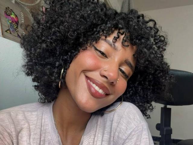 CinthiaWalker with curly black hair, smiling, wearing a gold nose ring and hoop earrings, in a light pink shirt, showing her teeth. Her brown skin is smooth.