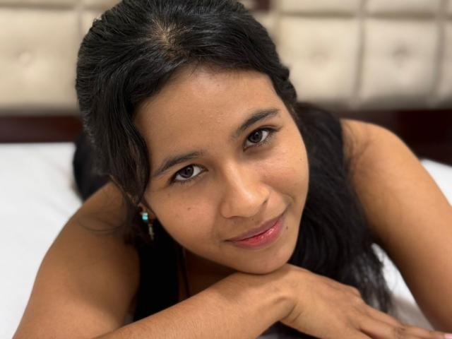 ArianaWright with dark hair, brown eyes, and a slight smile, looking at the camera, resting her chin on her hands, wearing a black top, on a bed.