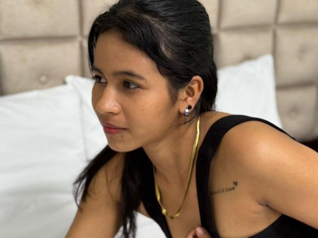 ArianaWright with dark hair, brown skin, small gold hoop earrings, black tank top, gold necklace, slight red lipstick, and tattoo on right shoulder, sitting on a bed.