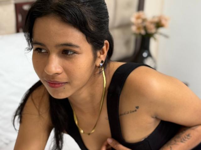 ArianaWright with dark hair, brown eyes, and tan skin, wearing a black tank top, gold necklace, and earrings, showing a tattoo on her right shoulder and a tattoo on her arm.