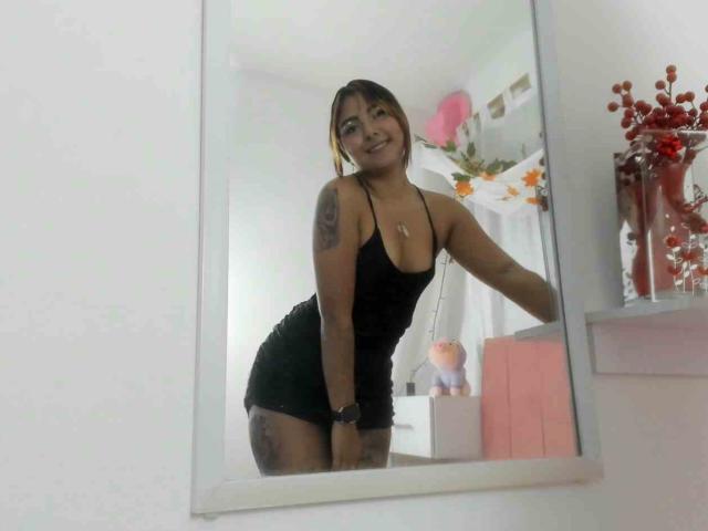VeronicaOnix with tan skin and tattoos, wearing a black, low-cut, tight dress, leaning in a mirror, smiling, showing her small, perky tits and ass. Mirror reflection.