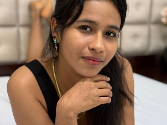 ArianaWright has medium brown skin, black hair, and is wearing a black top, with a gold necklace and teal earrings. She's resting her chin on her hand, smiling.