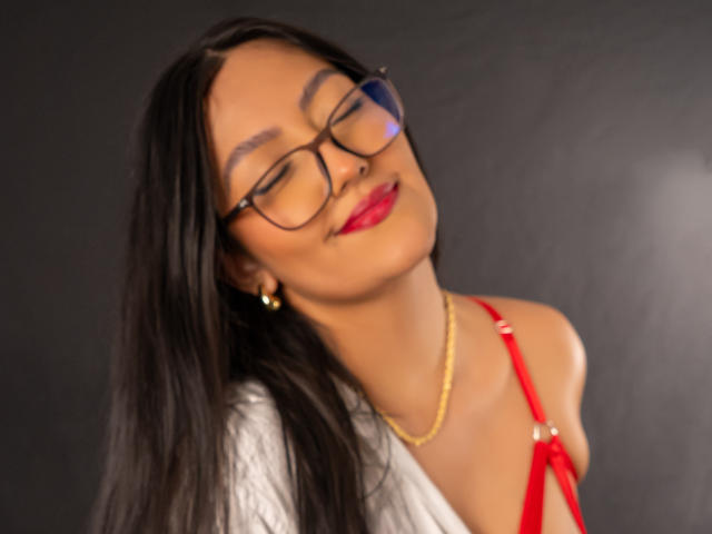 IsabellCollin with glasses, red lipstick, and a red bra, has her eyes closed and head tilted back, wearing a gold necklace against a dark background.
