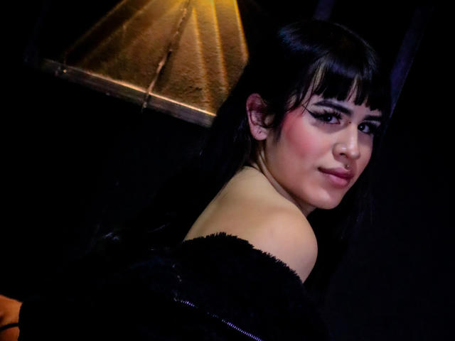 HannaMitsury with black hair and blunt bangs, wearing a black off-shoulder top, looking back with a subtle smile, nose ring, and pink blush on her fair skin.