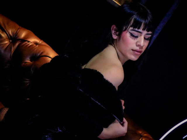 HannaMitsury with dark hair, bangs, and blue eyeliner, sits on a brown leather couch, black fur coat off her shoulders, showing her bare back and slightly turned face.