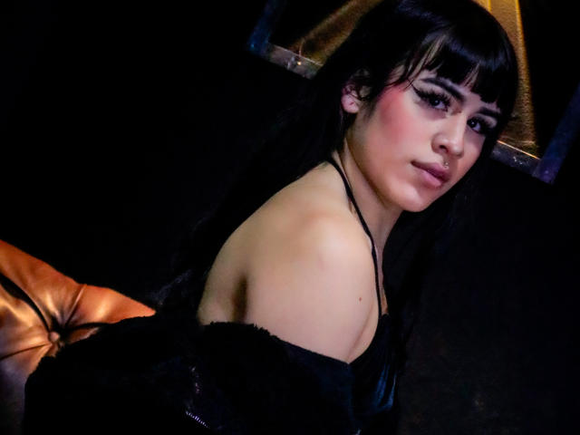 HannaMitsury with black hair, bangs, and heavy eyeliner, wearing a black off-shoulder top, sitting on a brown leather couch, looking over her shoulder with a slight smile.