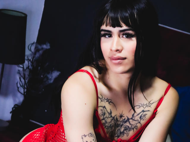 HannaMitsury with black hair, bangs, and red lingerie, showing tattoos on her chest and arms, leaning forward with a slight smile, looking directly at the camera.