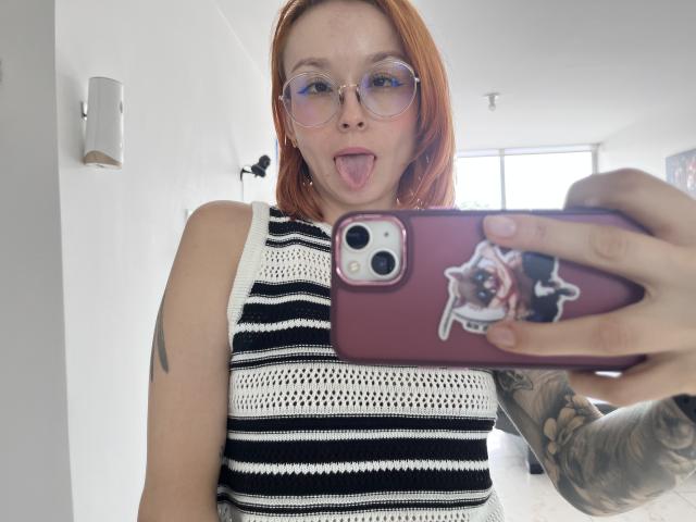 SarahRousen with red hair, glasses, tongue out, wearing a black and white striped sleeveless top, holding a pink phone with a cat sticker, tattoo on right arm.