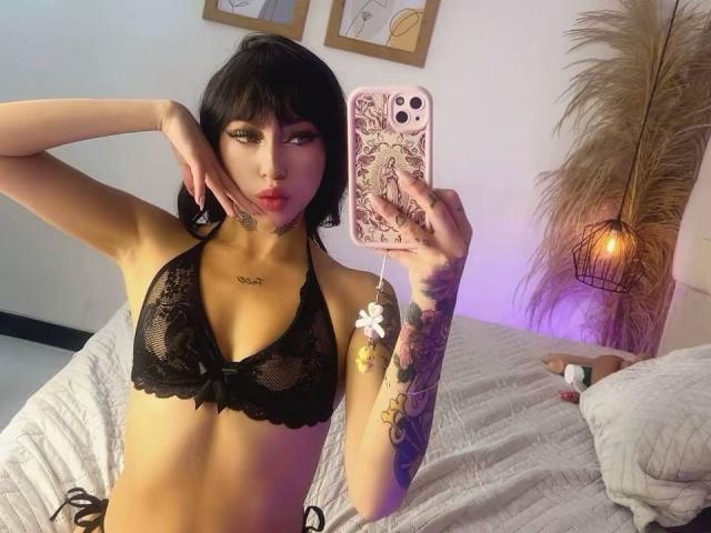 LunaBaster with black lace bra, tattoos, and short black hair, taking a selfie in a bedroom, showing off her small tits and flower tattoo on her right arm.