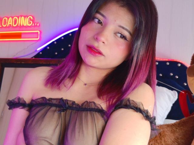 ShofiaWett with pink-tipped black hair, wearing a sheer black off-shoulder top, showing her tits, red lips, and a subtle necklace, looking seductively at the camera.