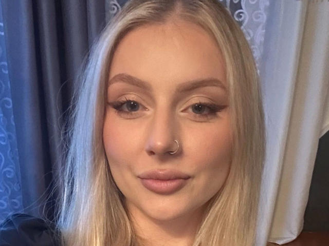 Close-up of MayaGeheim-hot, a blonde woman with straight, medium-length hair. She has a fair complexion, light makeup with defined eyebrows, long eyelashes, and subtle pink lipstick. Her nose is adorned with a small, silver nose ring. She looks directly at the camera with a neutral expression. The image is well-lit, highlighting her facial features. The background is blurred, with a mix of dark and light colors. The focus is on her face, showing her clear skin and slight smile.