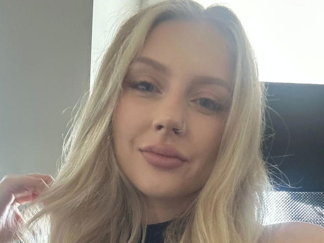 Close-up of MayaGeheim-hot with blonde, slightly wavy hair and light skin. She has a small nose ring in her right nostril and subtle, natural-looking makeup with light pink lipstick. Her eyebrows are thin and well-defined. She's looking directly at the camera with a slight, relaxed smile. The lighting is soft, highlighting her facial features. Her right hand is gently touching her hair. The image has a casual, candid feel.