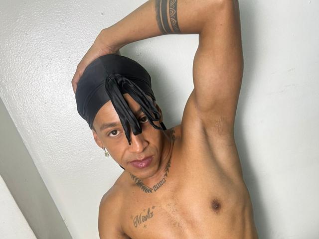 A photograph of HanselQueen, a muscular, dark-skinned man with a tattooed chest and visible tattoos on his arm. He wears a black headwrap with loose, hanging strands, and a small hoop earring in his left ear. His right arm is raised, hand resting on his head. His chest features a tattoo that reads "Blessed" and another tattoo below it. His expression is neutral, looking slightly to the left. The white wall behind him is plain and textured. The image is clear with sharp details, focusing on HanselQueen's upper body and face.