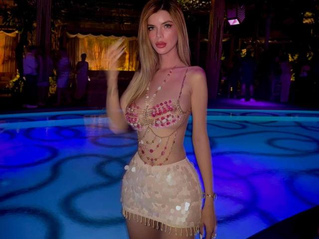 HotCumAdriana69x in a white lace dress, sitting on a pool edge, legs crossed, showing her ass, with red lipstick, in a nighttime pool party setting.