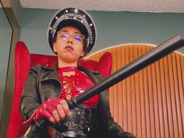 BarbaraLeythonn wearing a black leather jacket, red latex top with lace-up front, red gloves, and a black-and-white beaded cap, holding a black bat, looking down.