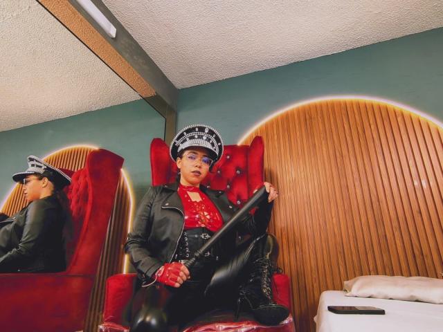 Barbara Leythonn, con una chaqueta de cuero negro, un corpiño de látex rojo y un sombrero, sosteniendo una porra negra, usando guantes rojos, sentada en una silla roja con paneles de madera..