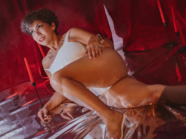 JuliaNewell with short curly brown hair, wearing white lace bra and panties, covering her pussy with her hand, standing against red curtain, looking sexy with open mouth.